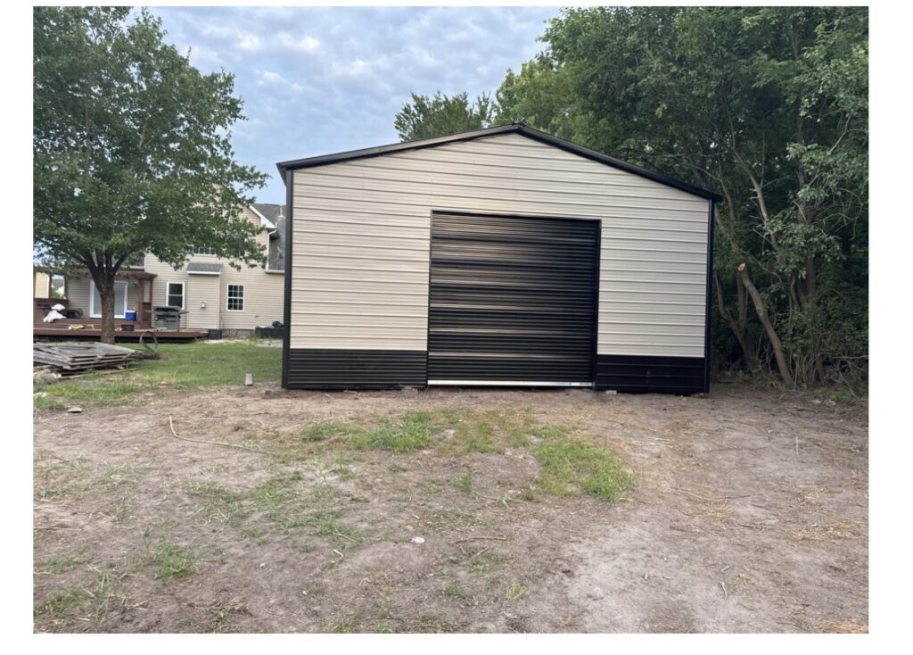 Tan metal garage with a black roll-up door on a gravel pad